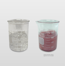 Two clear glass beakers on a light gray background, one filled with plain Versagel ME 750 lip gloss base and the other with a prepared red/pink lip gloss made with Versagel ME 750. A small note below the pigmented beaker says "tinted example shown, pigments not included."